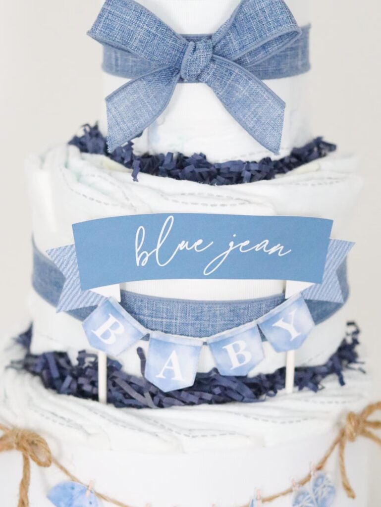 Tiered diaper cake decorated with blue denim ribbon, a large blue bow, and a mini banner for a blue jean baby shower theme.