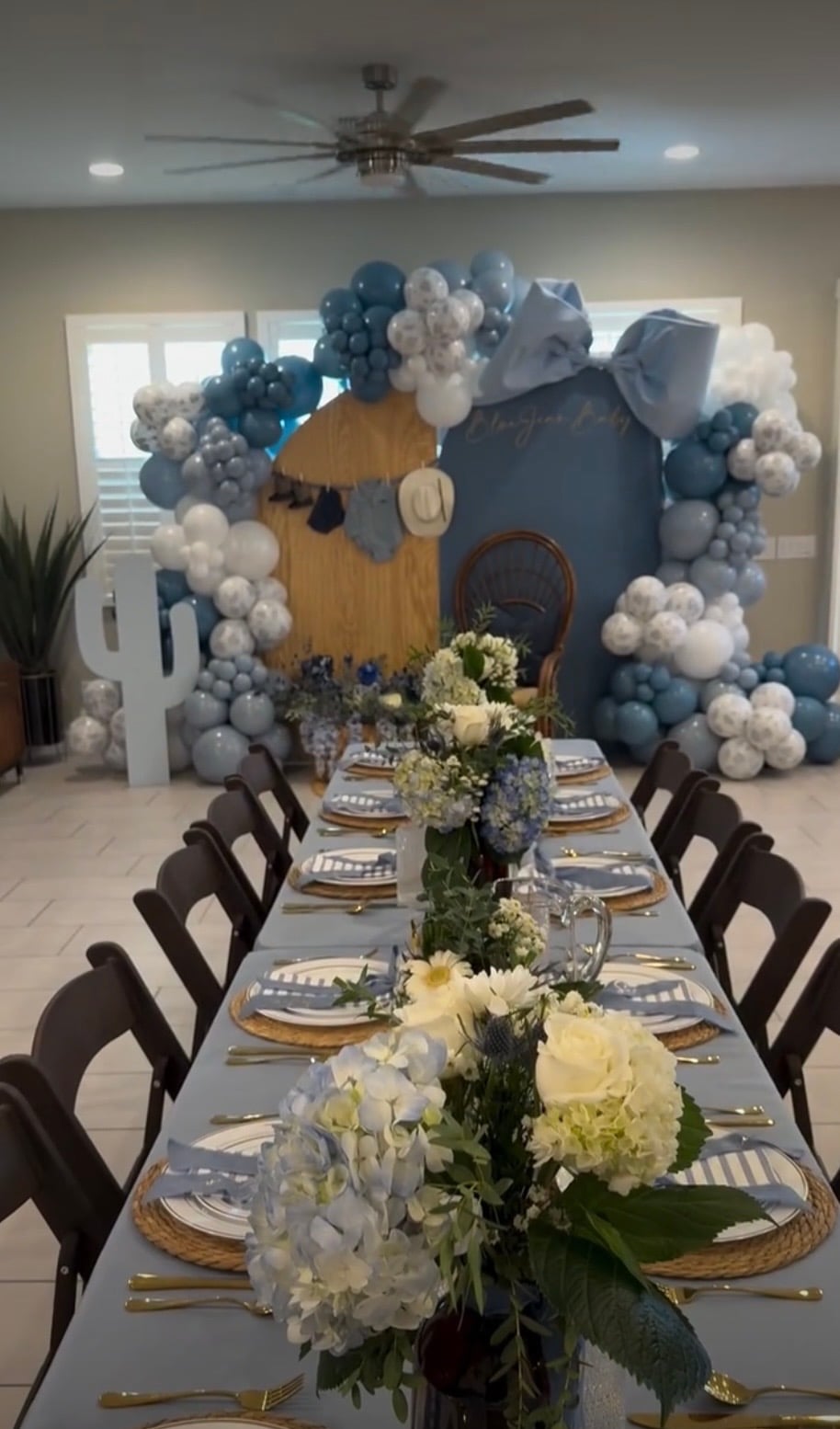 Blue Jean Baby shower decor with blue and white balloon garlands, an oversized blue bow, denim baby clothes, and a long table with blue and white floral centerpieces for a blue jean baby shower theme.