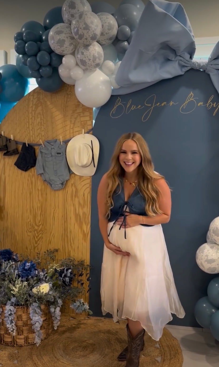 Pregnant woman wearing a denim top, white skirt, and cowboy boots in front of blue balloons, an oversized bow, and denim baby clothes for a blue jean baby shower theme.