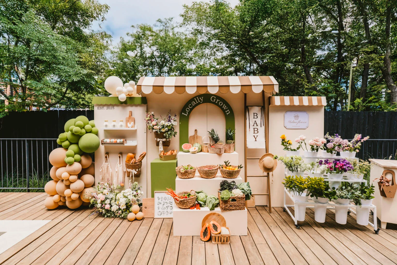 Locally grown baby shower market stand with fresh produce, flowers, woven baskets, greenery, and neutral balloon decor for a farmers market baby shower theme.