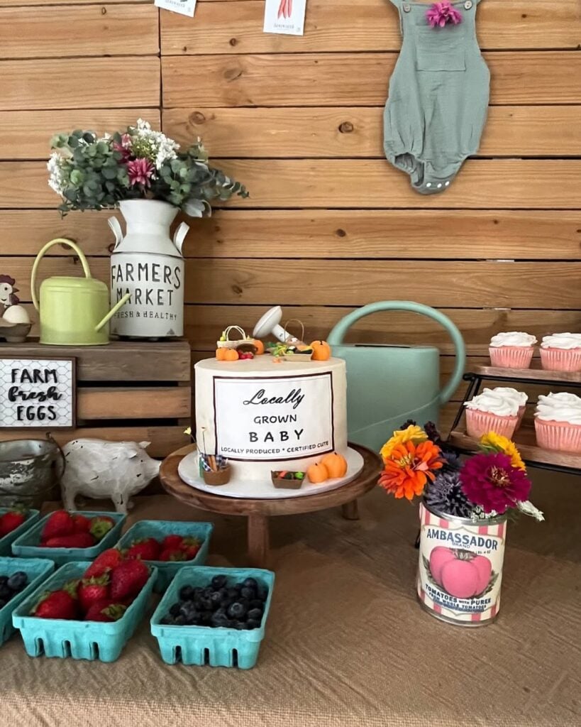 Locally grown baby shower dessert table with a farmers market cake, fresh berries, cupcakes, colorful flowers, watering cans, and rustic farm decor.