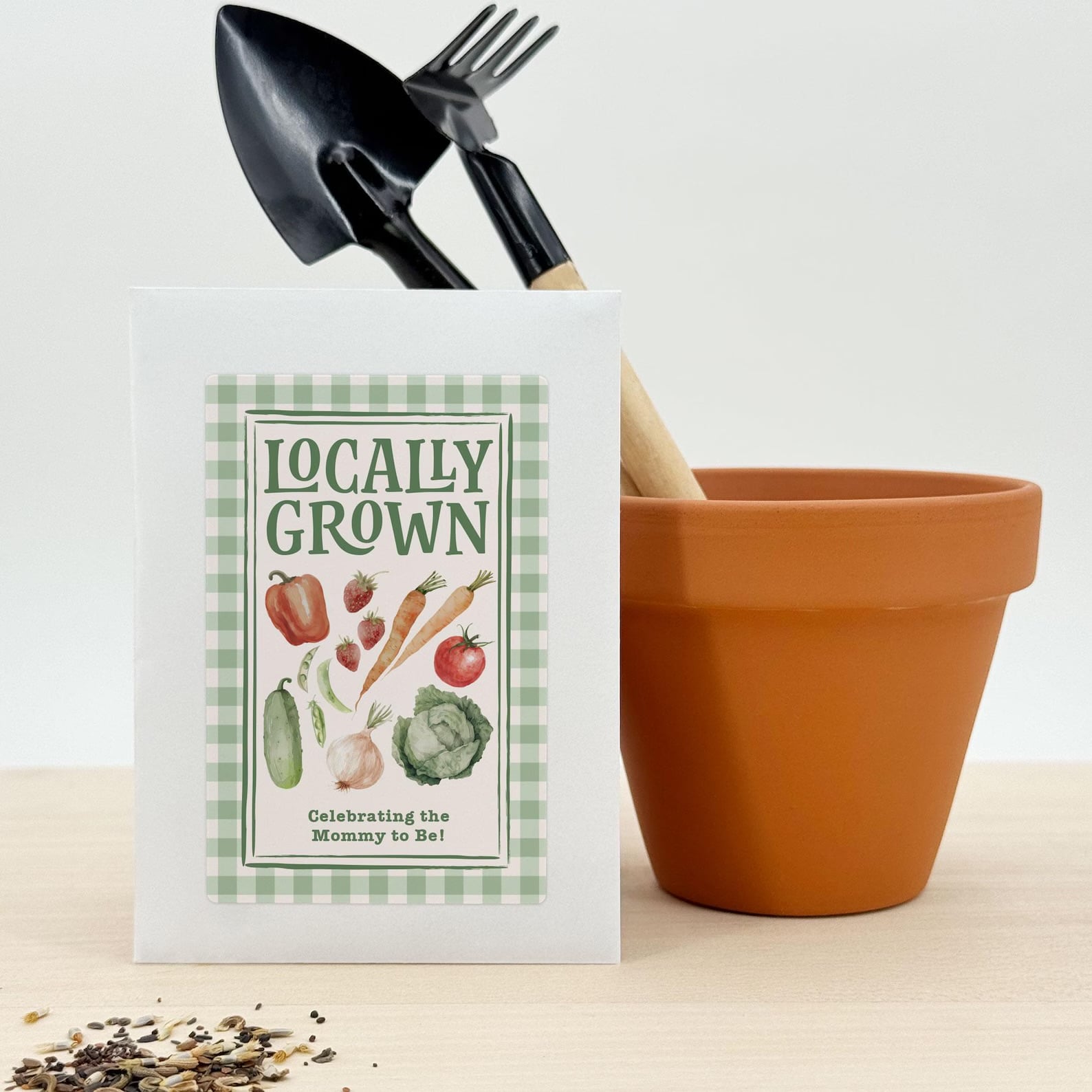 Locally grown baby shower favor bag decorated with green gingham and garden vegetables, displayed beside a terracotta pot and gardening tools.
