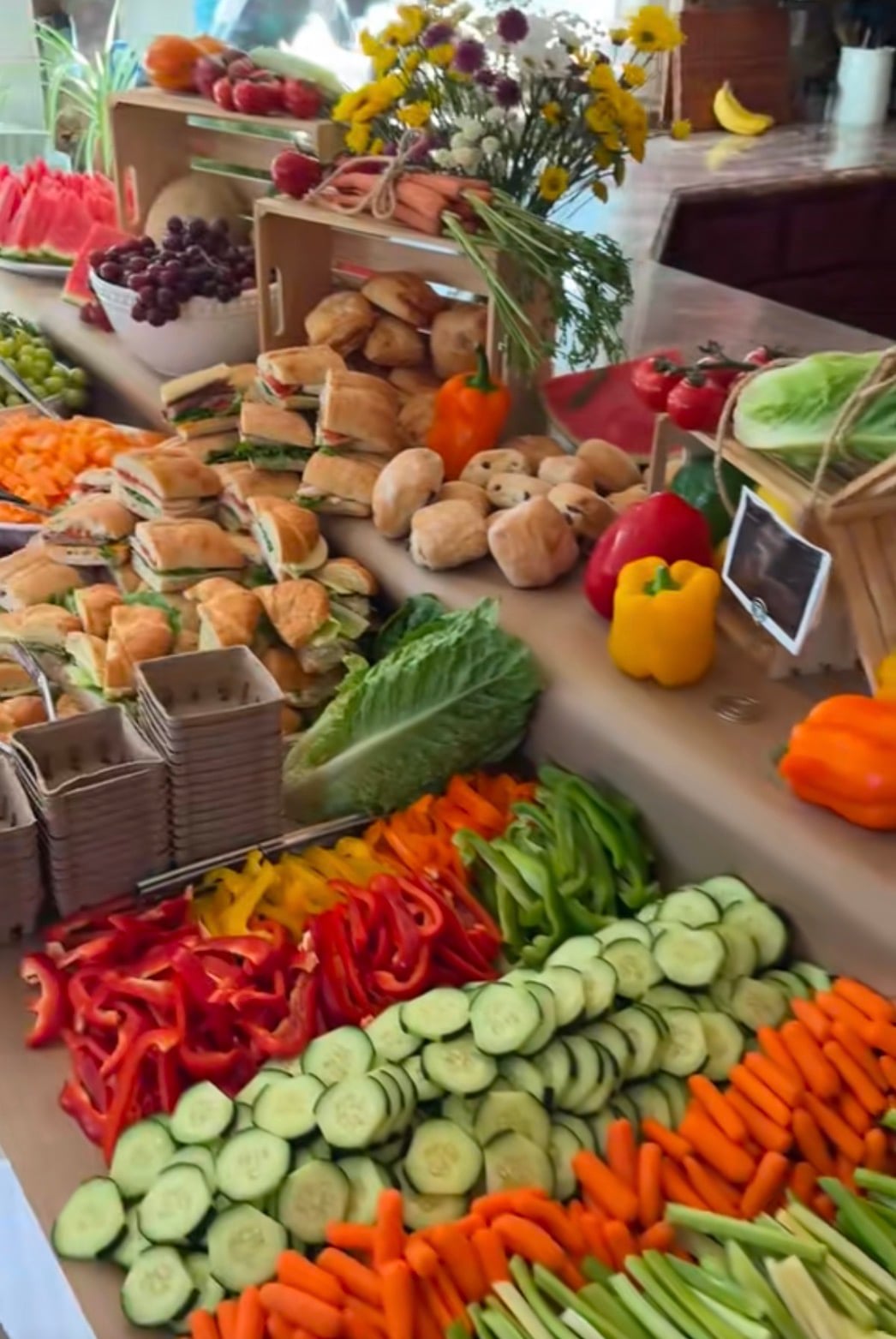 Locally grown baby shower food display with fresh vegetables, fruit, sandwiches, bread, and flowers arranged in wooden produce crates like a farmers market stand.