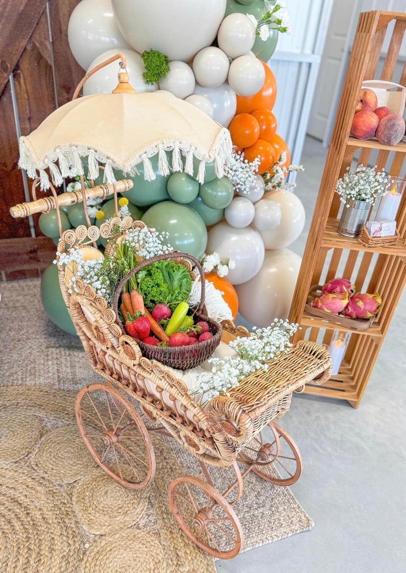 Locally grown baby shower decor with a vintage wicker stroller filled with fresh fruits, vegetables, and flowers in front of a sage green, orange, and cream balloon garland.
