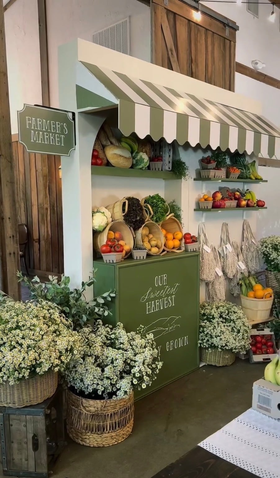 Locally grown baby shower farmers market display decorated with a sage green striped awning, baskets of fresh fruits and vegetables, market bags, and white flowers.