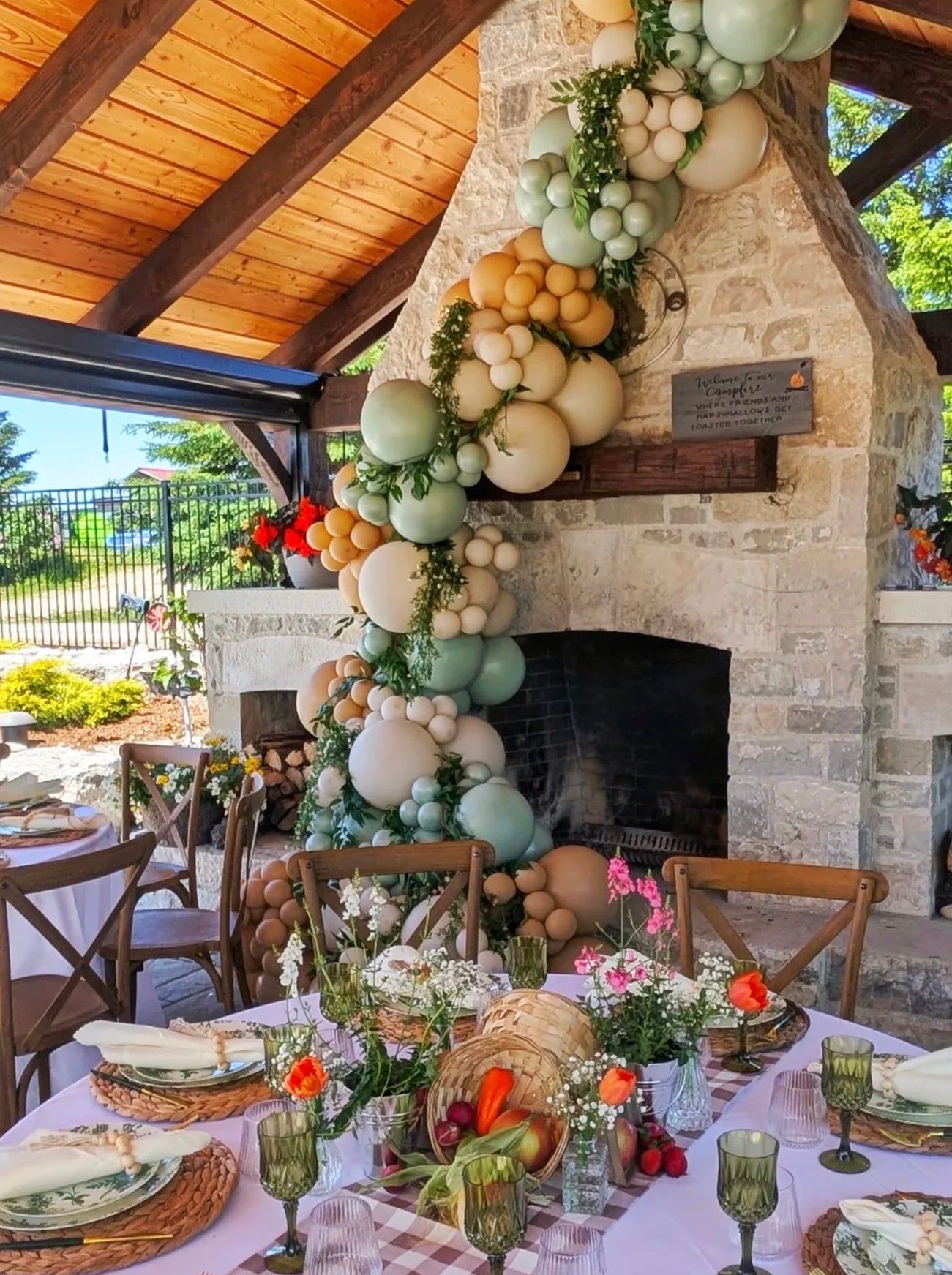 Locally grown baby shower table setting with a sage green, cream, and earthy orange balloon garland, fresh greenery, wildflowers, woven place settings, and garden vegetables.